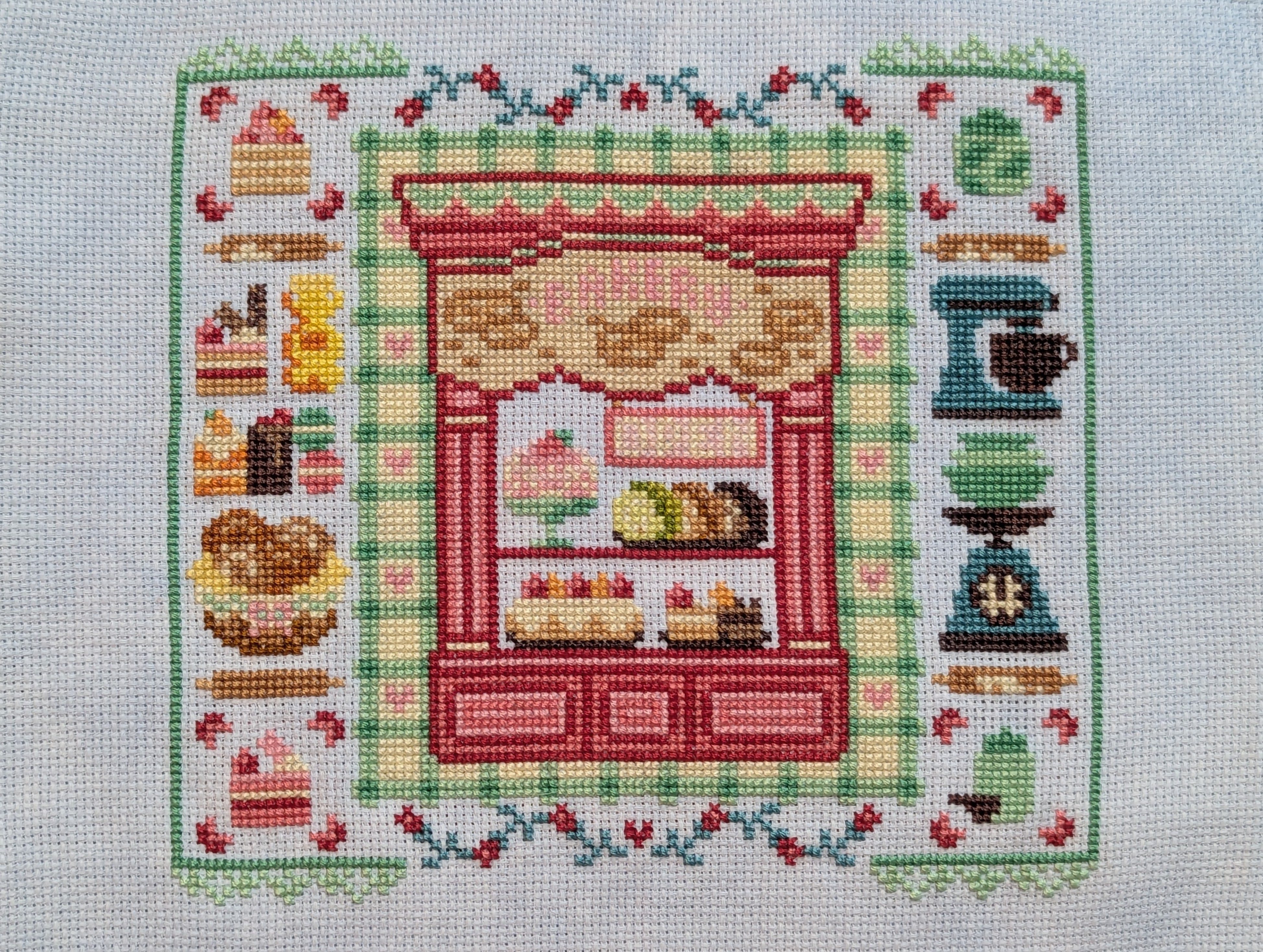 Cross stitch of a bakery storefront with baking tools (mixer, scale, rolling pins) and various baked goods surrounding it