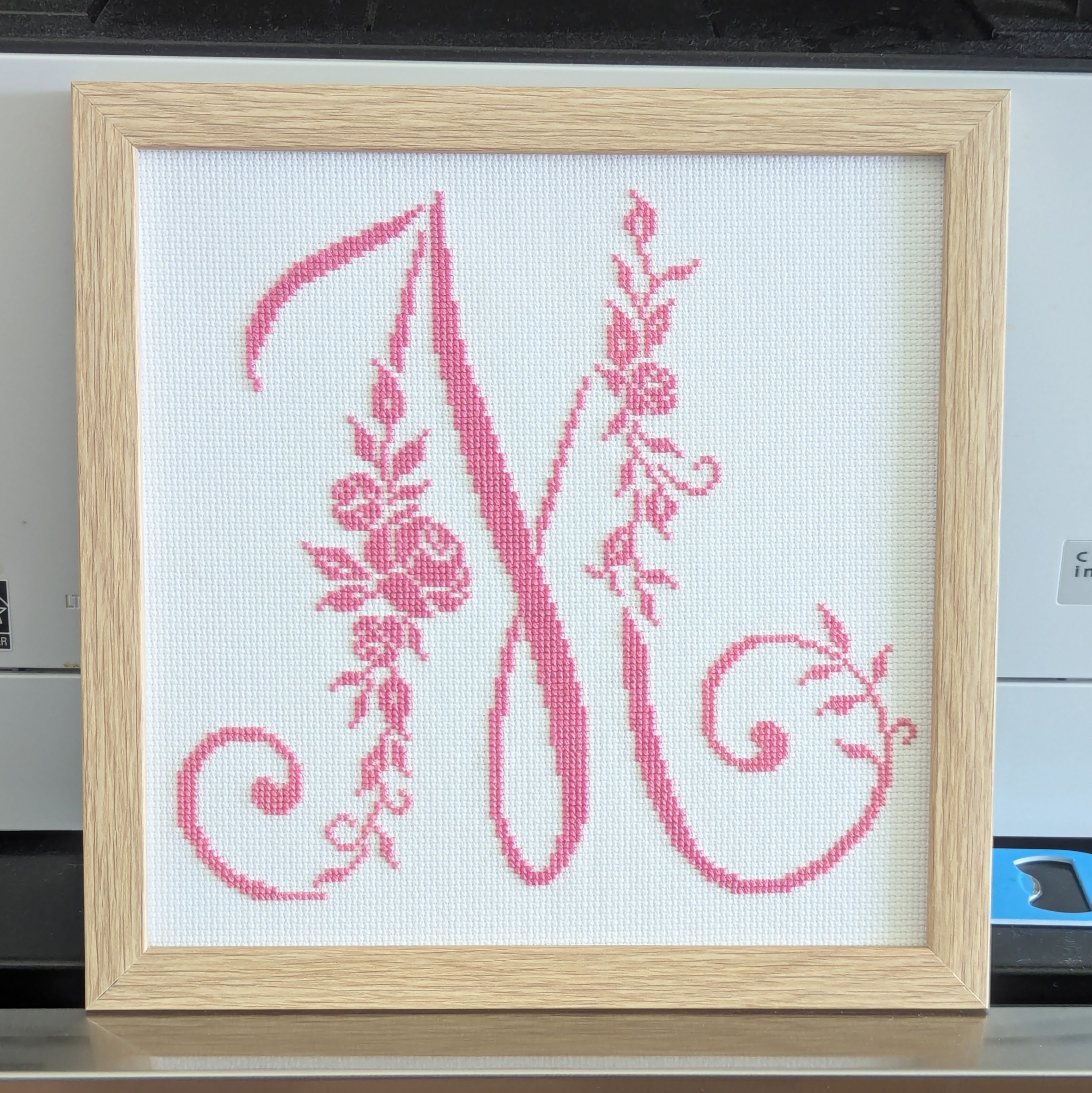 Cross stitch of a capital M in pink with roses and leaves making up part of the left and right verticals
