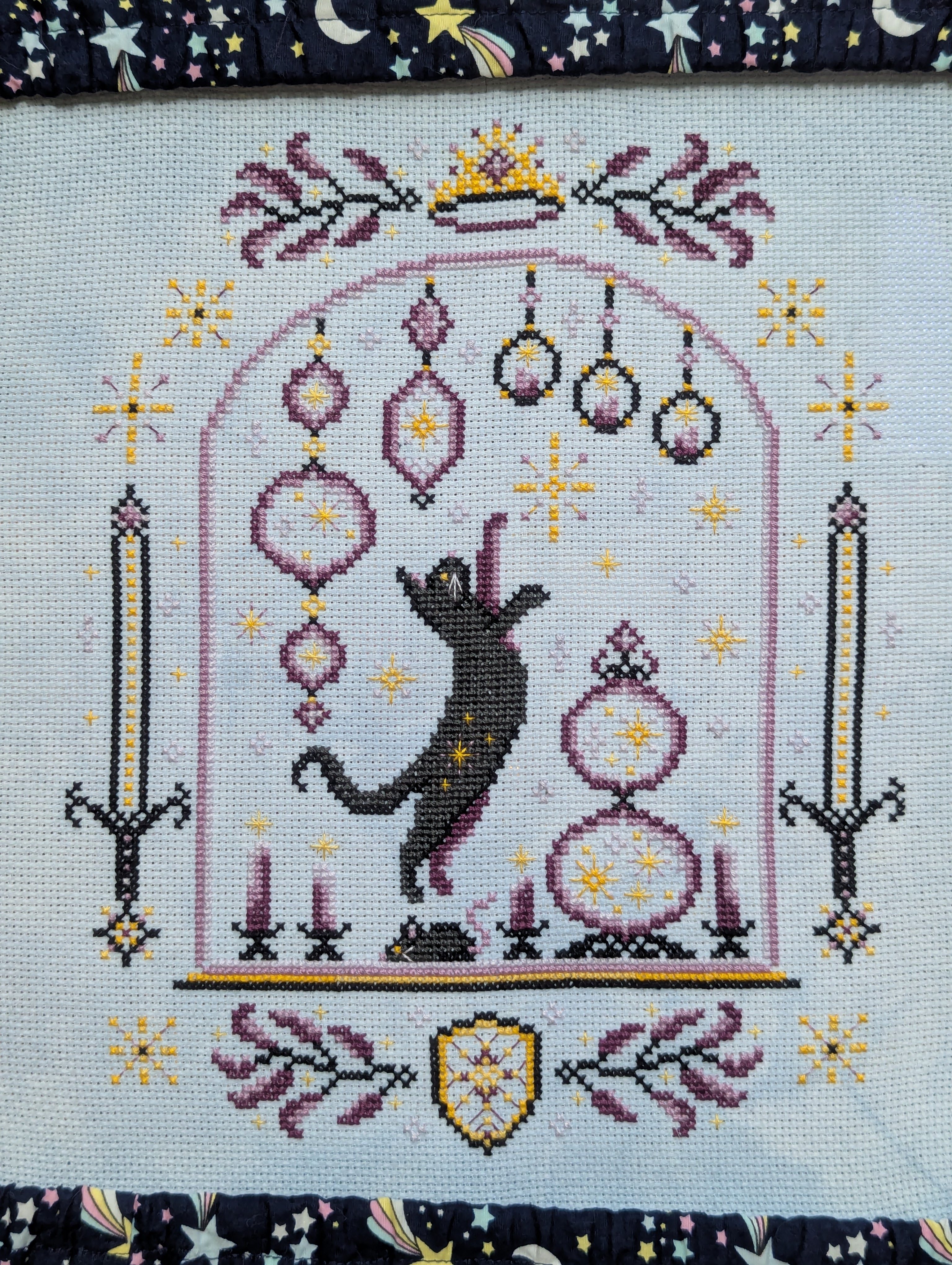 Cross stitch of an archway with a sword on each side. In the archway, a cat leaps with a paw stretched upward to bat at some hanging ornaments. There are candles and sparkles throughout.