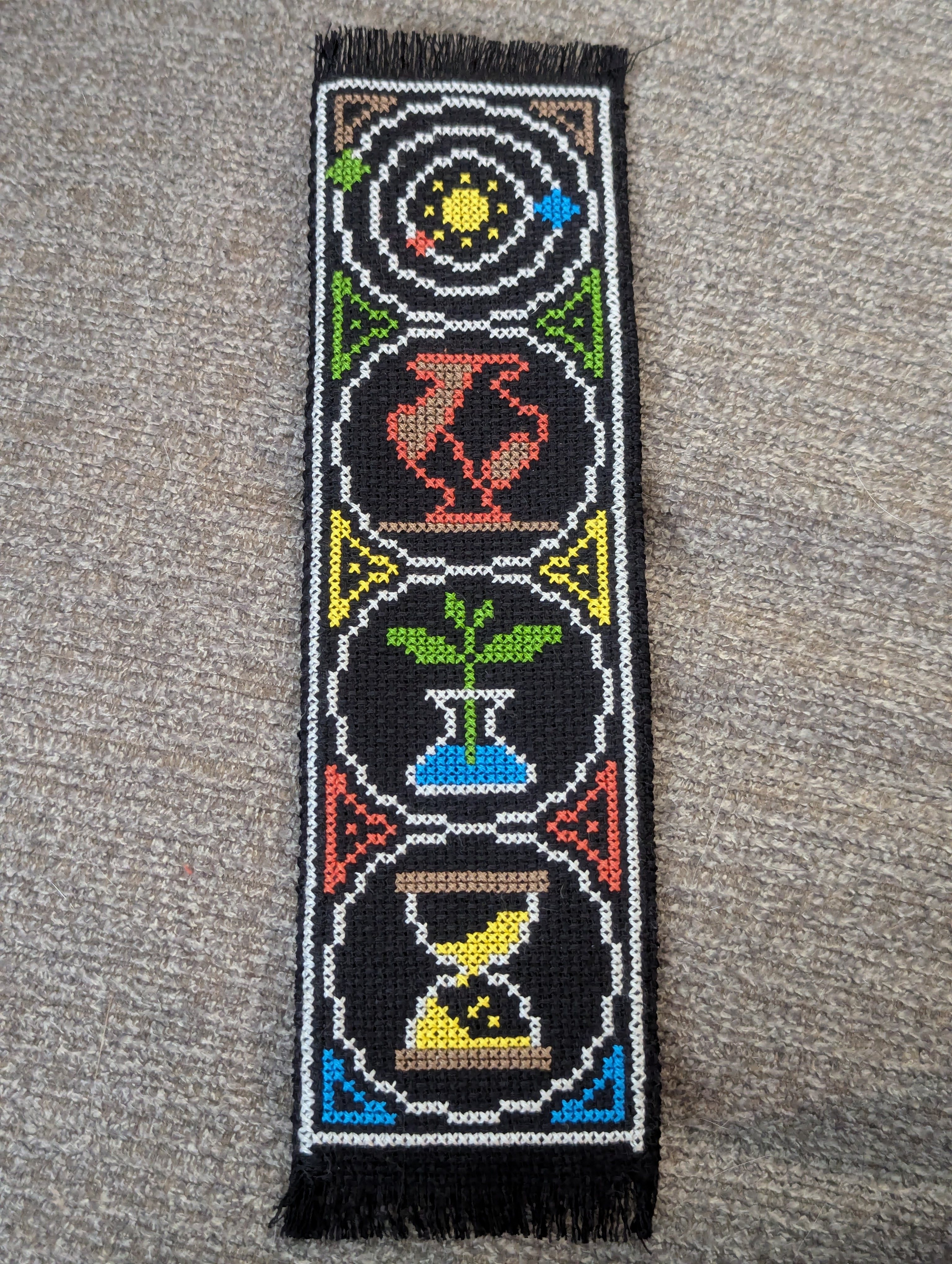 Cross stitch bookmark on black fabric depicting stylized images of a solar system, a cracked vase, a plant, and an hourglass